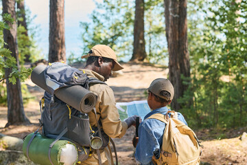 Back view of senior black man and his grandson with backpacks discussing map of local area while moving along forest