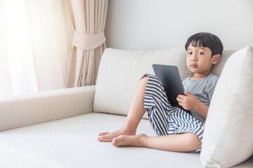 Fototapeta premium Adorable happy Asian boy wearing a gray shirt and blue-white striped shorts is having fun playing with his tablet on a cream sofa. looking at the mobile screen
