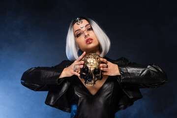 woman in leather jacket and halloween makeup posing with skull on dark and blue background.
