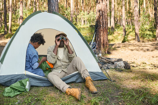Retired African American man in casualwear looking through binoculars while sitting in tent next to his grandson unpacking rucksack - Powered by Adobe
