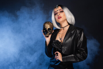 woman in witch halloween costume holding golden skull and black rose on dark background with blue smoke.