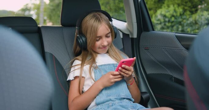 Small Stylish Girl Smiling Riding In Car, Listening To Music With Headphones On Smartphone In Backseat Of Car.