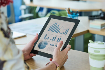 Hands of young female economist holding tablet with financial document on screen while sitting by workplace and analyzing data