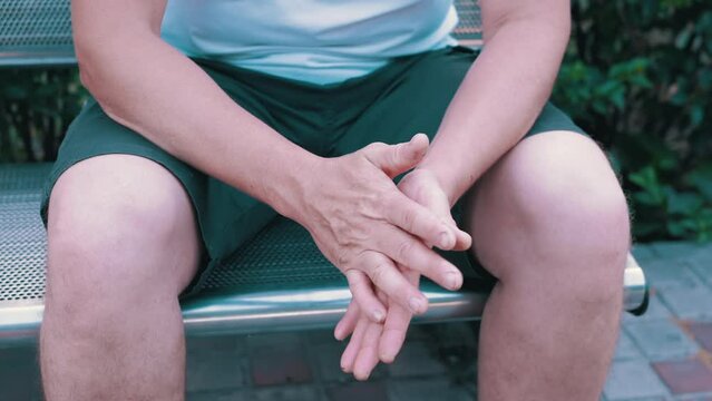 Male Rubbing Hands Sitting on a Bench in the Urban Park. A moment of waiting, excitement, anxiety, and emotional stress, trouble, nerves, and dread. Massage of hands, joints, fingers, skin. Outdoors.
