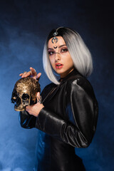 ash blonde woman in halloween makeup and leather jacket holding golden skull on black background with blue smoke.