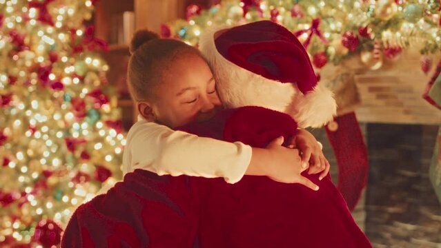 Waist up slowmo of happy little African American girl embracing Santa Claus next to beautifully decorated Christmas tree and fireplace