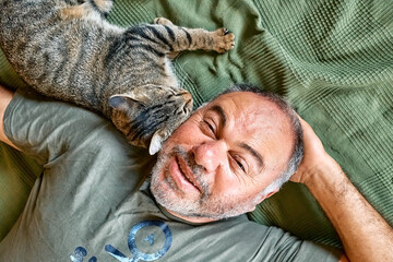 Top view of bearded middle-aged man lying on a bed and playing with his gray tabby cat....