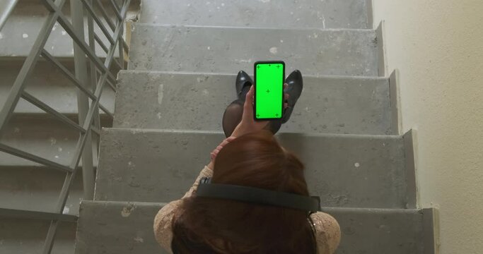 Caucasian young woman listens to music in headphones and looks at the smartphone screen. Staircase, concrete steps, top view. Concept of loneliness, kicked out of the house, returning after a party.