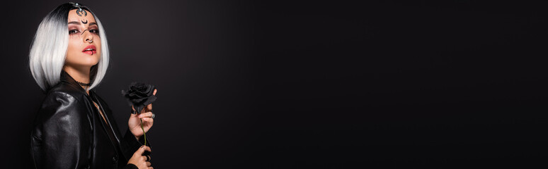 sexy woman in witch halloween costume holding dark rose isolated on black, banner.