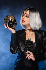 sexy woman in witch costume holding black rose and looking at golden skull on dark background with blue fog.