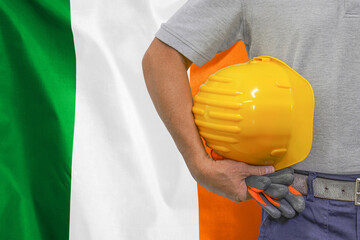 Close-up of hard hat holding by construction worker on Ireland flag background. Hand of worker with...