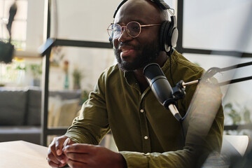 Young black man in headphones and eyeglasses talking in microphone while sitting by workplace and...