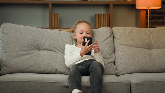 Active little kid girl jump on comfortable sofa in living room use modern gadget smartphone scroll pictures watch funny videos chatting with friends play mobile games break relaxation daytime at home