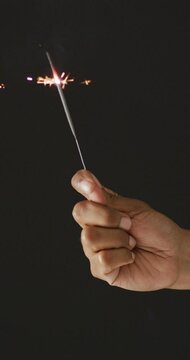 Vertical video of hand of african american person holding lit sparkler on black background