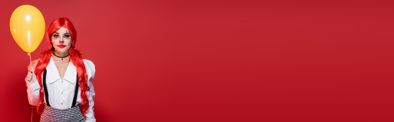 woman with bright colored ponytails and clown makeup holding yellow balloon isolated on red, banner.