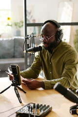 Young black man in headphones talking in microphone while reading his notes or plan of discussion in front of smartphone camera