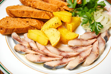 Pieces of pickled herring, potatoes, bread, onions and greens on a white plate