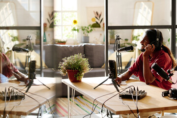 Young multi-ethnic man sitting by desk in front of microphone and talking to online audience while creating new audio file