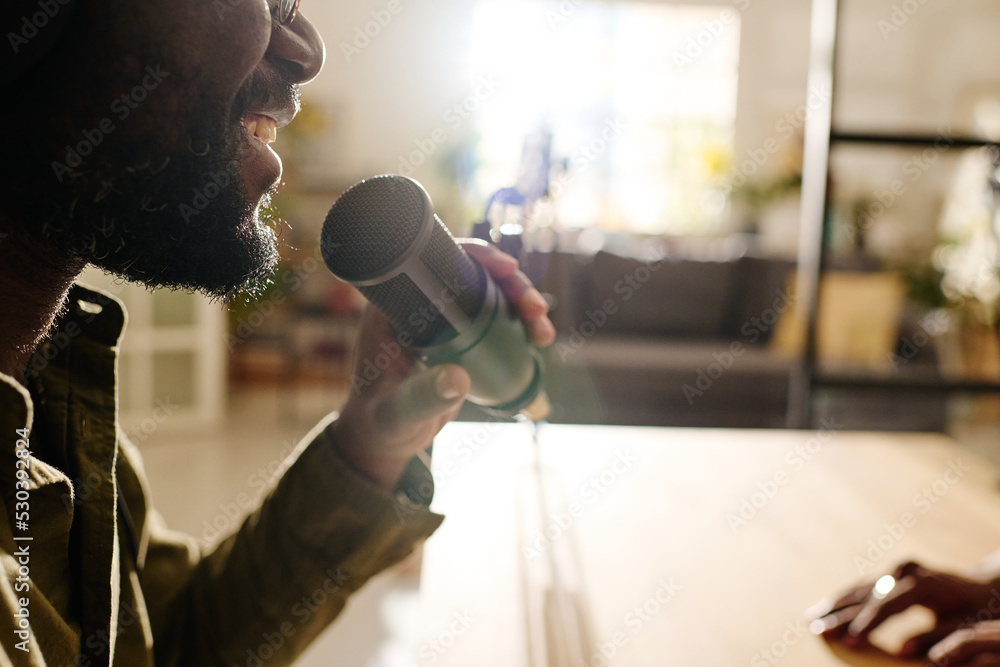 Wall mural Close-up of happy young bearded black man talking in microphone while sitting by workplace in front of camera and recording audio file