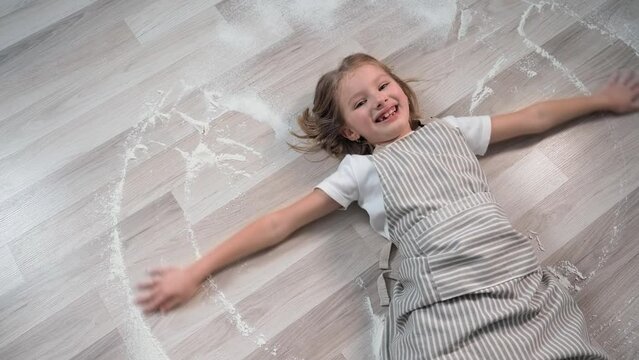 portrait of joyful little girl playing with flour lying on floor after cooking sweet buns at home in kitchen