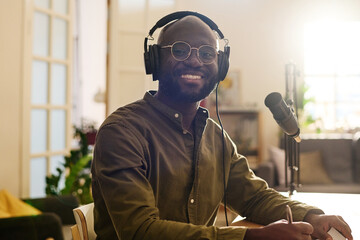 Young cheerful black man in headphones looking at camera while sitting by workplace in studio and writing down plan before recording audio