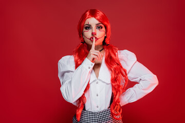 woman with clown style makeup showing hush sign while standing with hand on hip isolated on red.