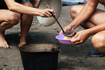 a family of tourists, a man and a woman pour into plates hot broth cooked outdoors on a fire in a...