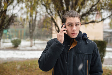 A young guy is still a teenager with a phone in his hand in winter, listening to what is being explained to him on the phone, the boy is already cold because he did not wear a hat