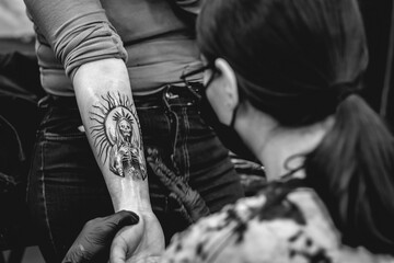 Detail of young tattoo artist girl with glasses and mask cleaning a new tattoo of 'La Santa Muerte' (Our Lady of Holy Death) in the arm of a woman (in black and white)