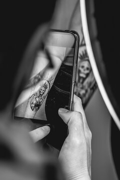 Detail of young tattoo artist girl with glasses and mask taking a photo with the phone of a new tattoo of 'La Santa Muerte' (Our Lady of Holy Death) in the arm of a woman (in black and white)
