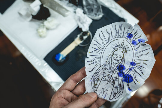Hand Holding The Design Of A New Tattoo Of 'La Santa Muerte' (Our Lady Of Holy Death) In Tracing Paper Over The Tattoo Artist Table With Tools And Materials