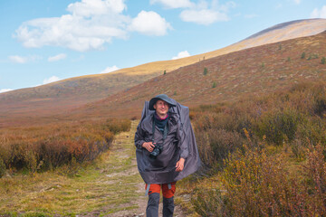 Alone traveler in raincoat with large backpack walks along hiking trail among fading autumn...