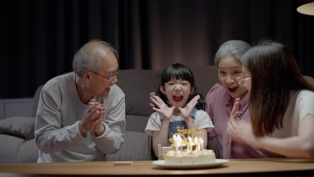 Asian happy family of little girl blowing out candles on cake. Celebrate birthday anniversary party with  Grandparents and mother on table at night in living room. Kid girl having happiness lifestyle.