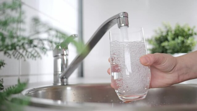 A stream of clean water drink flows into the glass. Woman holding a glass of water under running water from the tap in the kitchen.