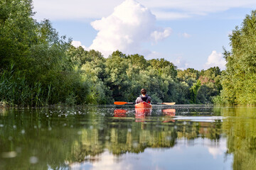 Obraz premium Rear view of man in red kayak. Kayaking on river near the shore with green trees against the blue sky and clouds in the background