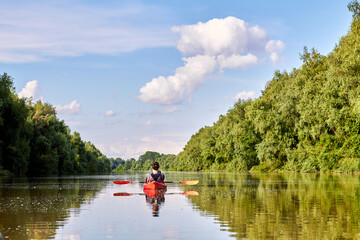 Rear view of man in red kayak. Kayaking on river near the shore with green trees against the blue sky and clouds in the background