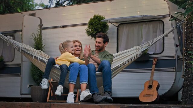 Caucasian family sitting in hammock in nature near camper van trailer at campsite weekend in camping outdoors cuddling relaxing talking chatting conversation give high five gesture of teamwork unity