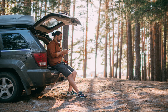A young man with a phone in his hands next to a car in a pine forest on the shore of a lake at sunset. A tourist with a smartphone and an SUV  on a forest road