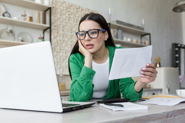 Cash loan. Upset and shocked woman in glasses holding bank bill about loan debt. Account cancellation. Sitting in the kitchen at the table with a laptop and documents.