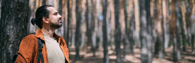 Portrait of a young man with a backpack on a walk in a pine forest on a sunny day. The guy breathes fresh air in a coniferous forest © Anna