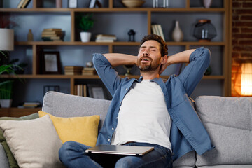 Handsome man working on laptop while sitting on sofa in stylish apartment. Relax after work. Relieved after work, put his hands behind his head and smiles.