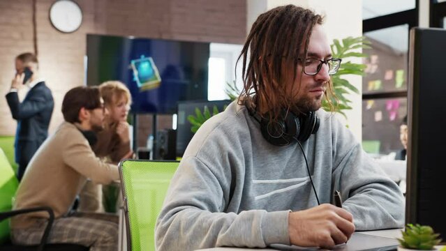 Caucasian male artist designer with dreadlocks and glasses draws on a tablet a 3D model for a computer game in the office of a project development studio. 