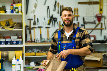 Handsome young carpenter sanding peace of wood and smiling while posing for camera