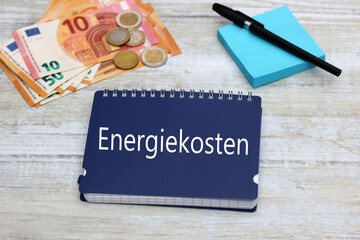 Blauer Notizblock mit dem Wort Energiekosten mit Eurobanknoten und Kugelschreiber.