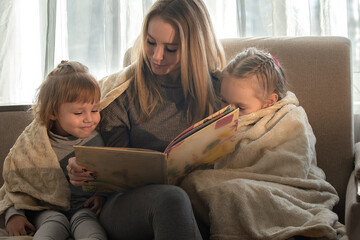 Mom at home on the sofa reads a book to her little daughters, they are wrapped in a blanket. Family developing leisure at home