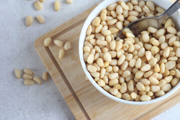 Peeled pine nuts lie in a plate on a wooden board. There is a spoon in the nuts. ,