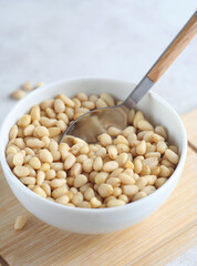 Peeled pine nuts lie in a plate on a wooden board. There is a spoon in the nuts
