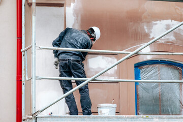 Renovation, restoration, refurbishment. Unrecognizable worker renovating wall of classical style building, standing on scaffolding. Construction worker prepares house facade wall for painting outdoors - obrazy, fototapety, plakaty