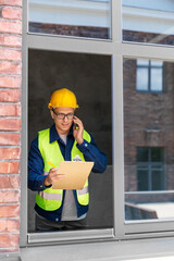 construction business and building concept - smiling male builder in helmet and safety west with clipboard calling on smartphone at window