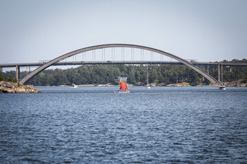 A sail boat with red sail is passing by a bridge. 